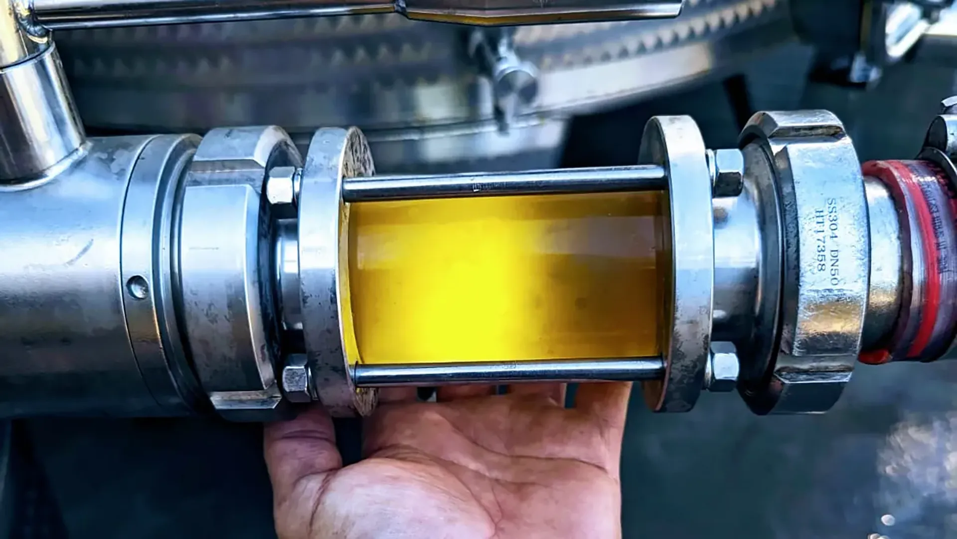 Tubo di acciaio inox con sezione trasparente contenente liquido giallo; mostra il flusso di produzione industriale.
