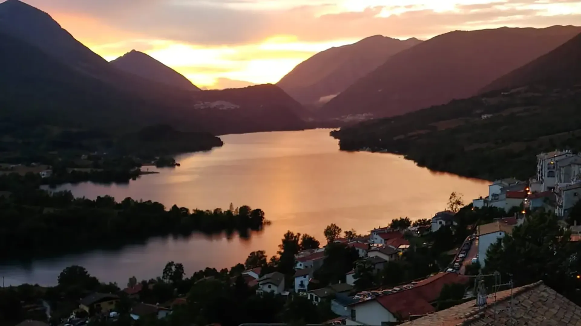 Tramonto sul lago con montagne scure e riflessi dorati sull'acqua, case in primo piano.