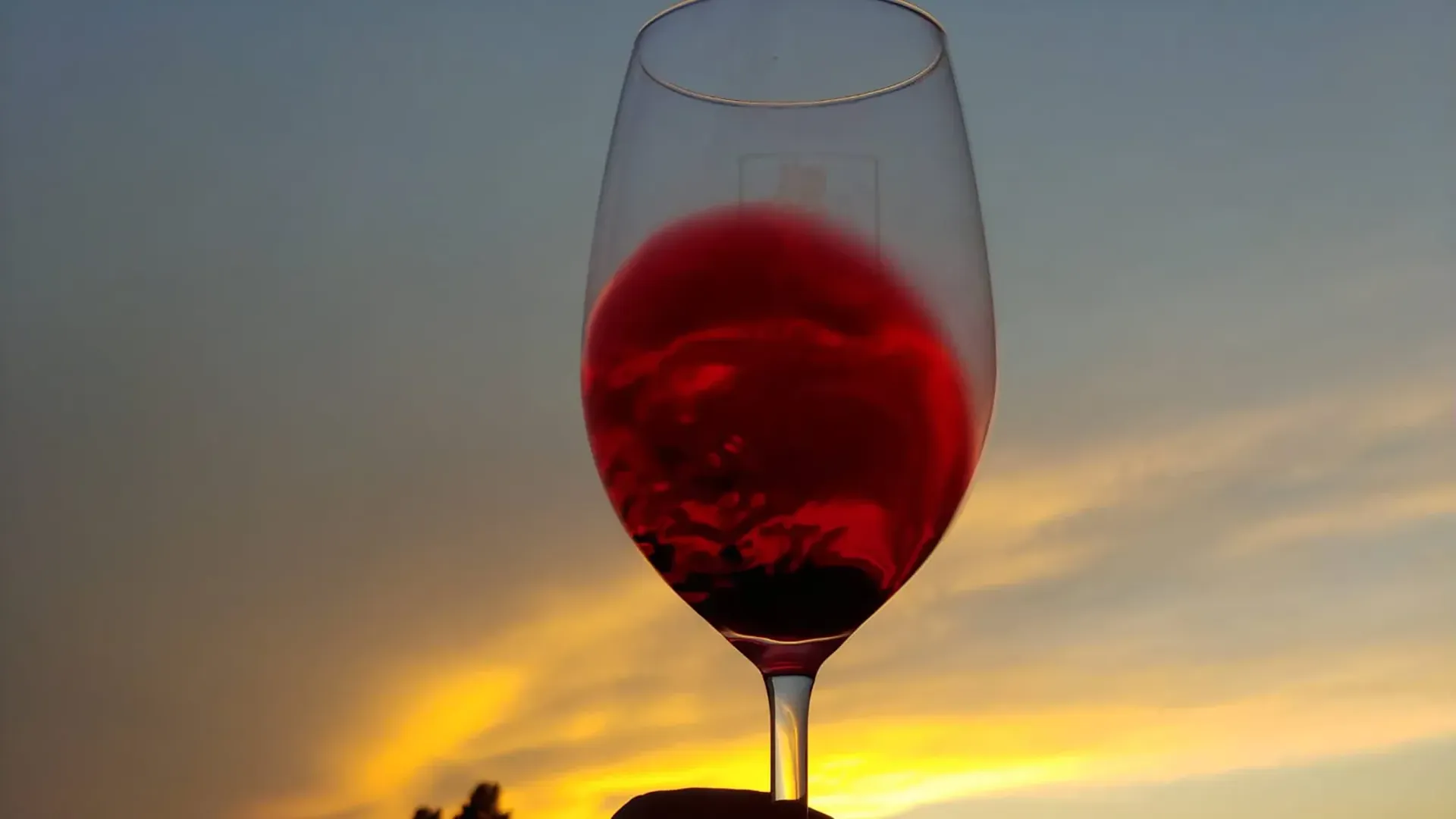 Calice di vino rosso con sfondo di tramonto dorato e cielo sereno.