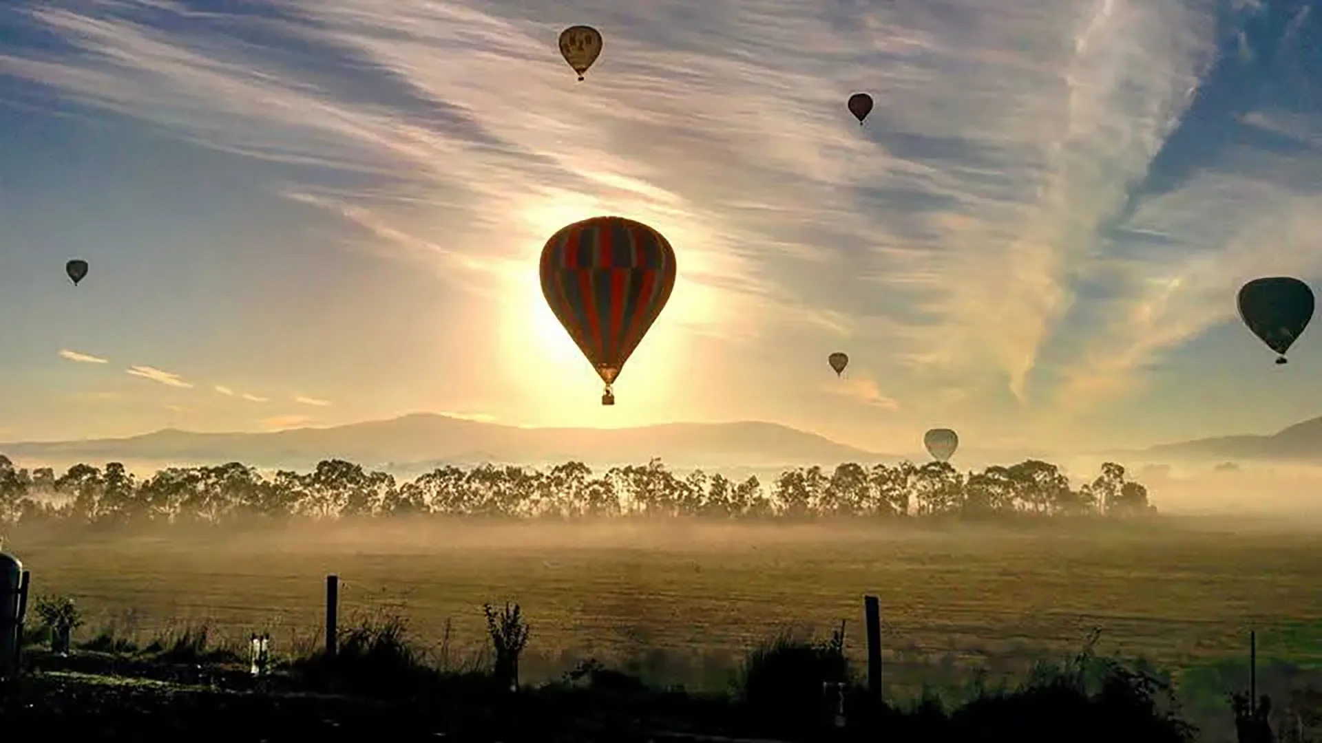Mongolfiere che sorvolano un paesaggio nebbioso all'alba, con il sole che splende dietro le colline.